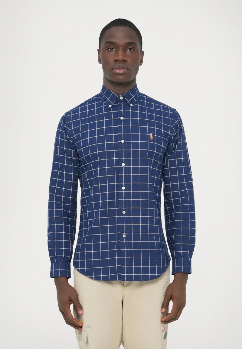 Temno modra srajca z dolgimi rokavi in gumbi, okrašena z belo mrežasto vzorcem in majhnim vezenim logotipom na prsih. Izdelana iz tkanine.