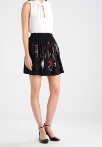 Žena v bezramenném bílém topu s výstřihem ve tvaru klíče, černé květované sukni a černých sandálech na kotníku, stojící na bílém pozadí.