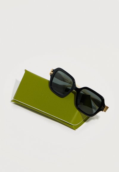 Lunettes de soleil à monture carrée noire avec verres foncés reposant sur un étui rectangulaire vert à bordure blanche, posées sur une surface blanche.