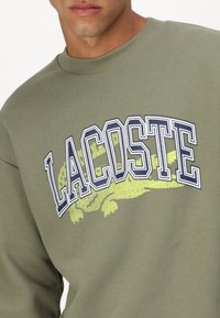 Sudadera verde oliva con un gran logo en blanco y marino de "LACOSTE" y un gráfico estilizado de un cocodrilo amarillo debajo del texto.