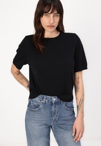 Schwarzer Strickpullover mit kurzen Ärmeln und Rundhalsausschnitt, kombiniert mit hochgeschnittenen Jeans aus Denim, die subtile rosa Verzierungen aufweisen.