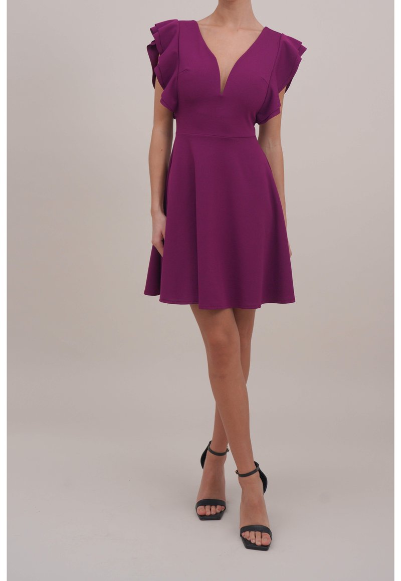 Femme portant une robe prune longueur genou avec des manches courtes à volants et un décolleté en V profond, associée à des talons noirs à bride à la cheville.