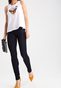 Top blanco sin mangas con un gráfico de mariposa; combinado con jeans oscuros ajustados y tacones amarillos de punta, sosteniendo un bolso de mano negro.