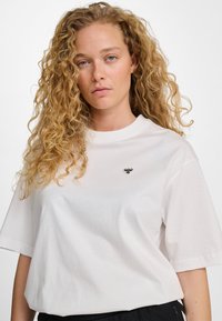 Biała bawełniana koszulka z okrągłym dekoltem i krótkimi rękawami. Posiada małe czarne haftowane logo na piersi oraz luźny krój.