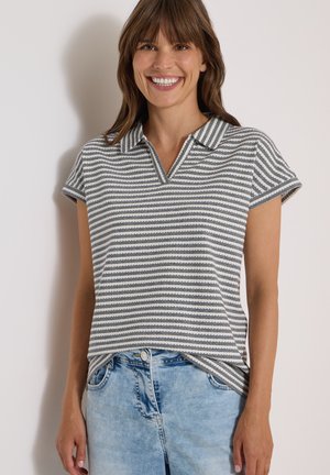 Lächelnde Frau, die ein grau-weiß gestreiftes Kurzarm-Poloshirt und hellblaue Jeans vor einem einfarbigen Hintergrund trägt.