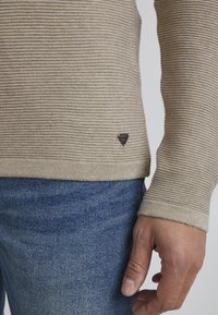 Krém színű kötött pulóver texturált, bordázott mintával, lekerekített aljjal és egy kis fekete logó címkével az alján, bal oldalon. Kék farmernadrággal viselve.