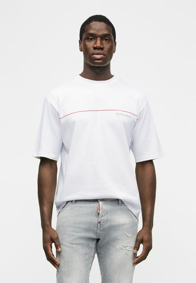 Άνδρας που φοράει λευκό μπλουζάκι με κόκκινη γραμμή και ανοιχτό γκρι ξεθωριασμένα τζιν, στέκεται μπροστά σε απλό λευκό φόντο.