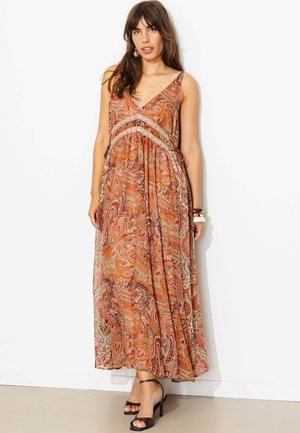 Femme portant une robe longue sans manches orange à motif cachemire avec bordure en dentelle, des sandales noires à talons hauts et des bracelets, debout contre un mur blanc.