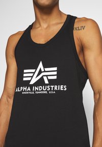 Schwarzes Trägershirt aus Baumwolle, mit einem weißen gedruckten Logo mit stilisiertem "A" und dem Text "ALPHA INDUSTRIES" auf der Vorderseite.