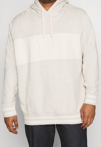 Person, der einen cremefarbenen und beigen Hoodie von Levi Strauss & Co. mit Fronttasche und Kordelzügen trägt, kombiniert mit dunkelblauen Jeans.