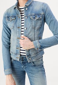 Lichtblauwe denimjack met een getailleerd ontwerp, twee borstzakken, metalen knopen en een klassieke kraag. Draagt over een gestreept shirt.