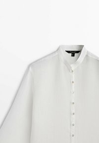 Chemise blanche à manches longues avec boutons, col mandarin, tissu léger et étiquette de marque visible à l’intérieur du col.