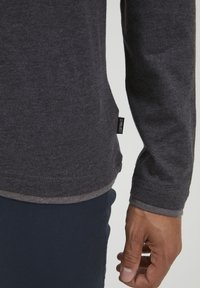Donkergrijze langemouwen shirt met een afgeronde zoom, voorzien van een klein zijlabel en een zachte textuur. Gekleed met marineblauwe broek, waarbij de handplaatsing zichtbaar is.