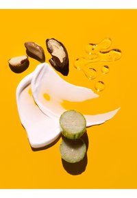 Trzy połówki orzecha brazylijskiego, dwie plastry selera, biały kremowy ślad i pięć kropli oleju na jasnym żółtym tle.