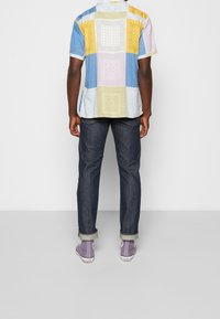 Korte mouwen shirt met een patchwork ontwerp in blauw, geel en pastelkleuren. Draagt met een donkere spijkerbroek en paarse sneakers.