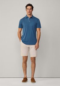 Blå kortärmad pikétröja med krage och bröstficka, tillsammans med ljusbeige shorts och bruna mockasiner.