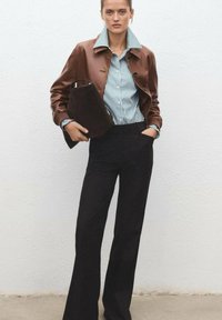 Brown leather cropped jacket over a blue and white striped shirt, paired with black flared trousers. She holds a dark clutch purse.