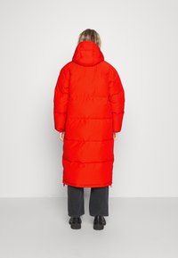 Rode lange pufferjas met een capuchon, voorzien van een gewatteerd ontwerp en geribde accenten. Het kledingstuk reikt tot halverwege het scheenbeen en is gemaakt van een gladde stof.