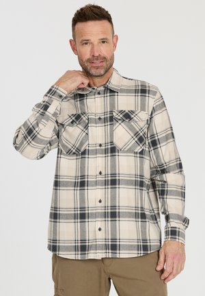 Mann, der den Kragen eines beige-grau karierten Hemds mit Brusttaschen anpasst, trägt dazu beige Hosen vor einfarbigem Hintergrund.