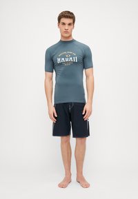 Herre kortermet rash guard i skiferblå med "Hawaii" trykk. Kombinert med svarte badebukser med hvite detaljer.