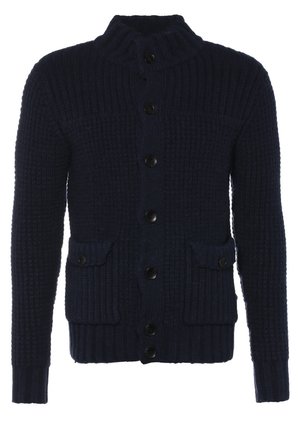 Uomo che cammina indossando un cardigan nero, una felpa grigia, pantaloni neri, sneaker nere e borsa grigia a tracolla su sfondo semplice.
