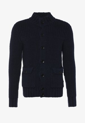 Homme marchant portant un cardigan noir, un sweat-shirt gris, un pantalon noir, des baskets noires et un sac bandoulière gris sur un fond uni.
