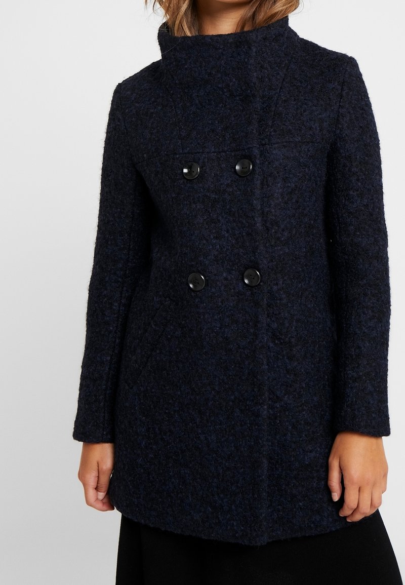 Cappotto in misto lana blu navy con colletto alto, chiusura doppio petto e due tasche frontali. Tessuto testurizzato con una finitura morbida.