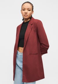 Νεαρή γυναίκα με πλεξούδες, φορούντας μακρύ μπορντό σακάκι, μαύρο κοντό τοπ και ανοιχτό μπλε τζιν ψηλόμεσο, με απλό φόντο.