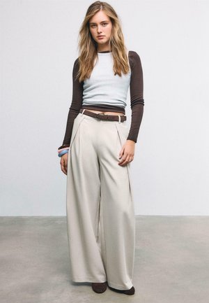 Jeune femme aux longs cheveux blonds portant des hauts marron et blancs à manches longues superposés, un pantalon large beige, une ceinture marron et des bracelets, debout à l'intérieur.