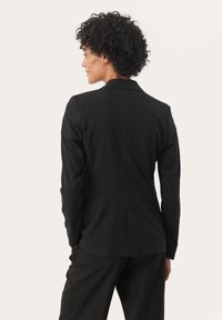 Blazer noir à la coupe ajustée, avec des revers à cran et deux fentes dans le dos. Texture lisse et manches longues, associé à un pantalon assorti.