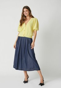 Kollane ribaga kootud kardigan lühikeste puhv-särkidega, mis on kombineeritud tumedate teksariidest plisseeritud midi-seelikutega. Mustad terava ninaotsaga kingad lõpetavad komplekti.