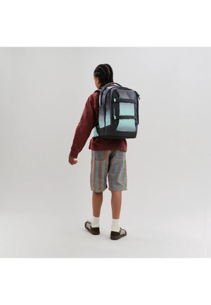 Kind mit geflochtenem Haar trägt einen großen schwarzen und hellblauen Rucksack, gestreifte Shorts, eine rote Jacke, weiße Socken und schwarze Turnschuhe, schaut von hinten.