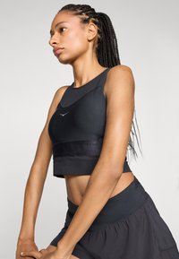 Čierny športový crop top s hladkým povrchom, so priliehavým strihom, širokým pásom a minimalistickým logom. Kombinovaný s čiernymi šortkami.