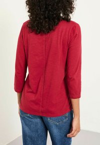 Rotes Longsleeve-Oberteil mit rundem Ausschnitt und Nahtdetails am Rücken, kombiniert mit blauen Jeans. Weicher Stoff und lockere Passform.