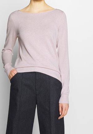 Mujer con suéter de manga larga color rosa claro metido ligeramente en pantalones anchos de pierna ancha, oscuros y de cintura alta, con una mano en el bolsillo.