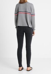 Šedý pletený svetr s červenými a námořnickými vodorovnými pruhy na rukávech, v kombinaci s černými skinny džínami a černými sandály. Pohled zád.