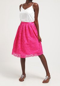 Kobieta ubrana w białą bezrękawnikową bluzkę oraz jasnoróżową koronkową spódnicę midi i srebrne sandały, stoi ze lekko zgiętą jedną nogą.