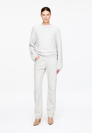 Kvinde iført lysegrå, blød sweater, elfenbensfarvede bukser og beige spidse hæle, stående med den ene hånd i lommen på hvid baggrund.