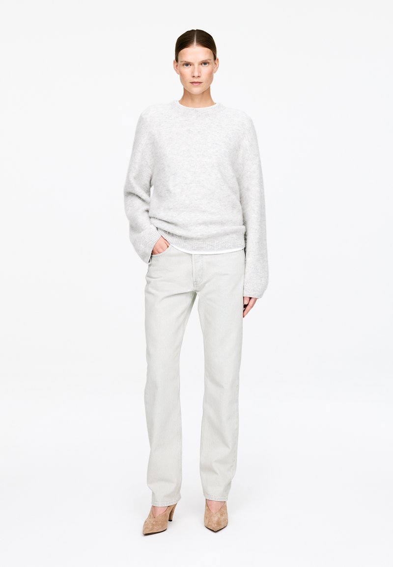 Femme portant un pull gris clair pelucheux, un pantalon blanc cassé et des talons beiges pointus, debout avec une main dans la poche sur fond blanc.