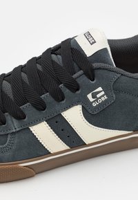 Zbliżenie na czarno-białe sneakersy marki Globe z czarnymi sznurowadłami, zamszową teksturą i brązową gumową podeszwą.