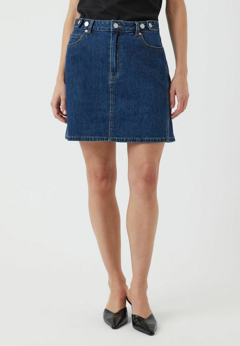 Tummansininen farkkuhame. Siinä on tiukka vyötärö, viisi taskua, näkyvää ommelta ja etupainikekiinnitys. Sileä pinta.