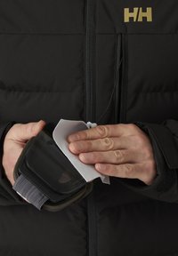 Schwarze Skibrille mit grauem Band, die mit einem weißen Tuch gereinigt wird, gehalten in einer Hand vor einem Hintergrund aus einer schwarzen, gepolsterten Jacke.