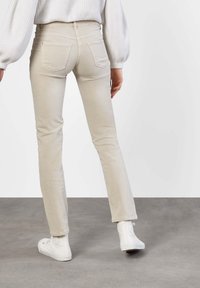 Pantalones ajustados beige con una textura suave, dos bolsillos traseros, combinados con zapatillas altas blancas sobre un suelo gris.