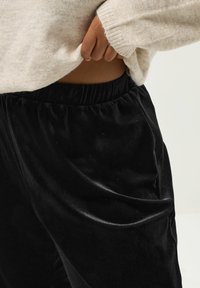 Svarta sammetbyxor med elastisk midja. Händerna framstoppade i en beige tröja med ribbade ärmslut, som visar upp en slät textur och mjukt material.