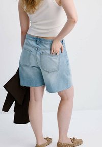 Person wearing light blue denim shorts and white tank top, holding a dark jacket in left hand, right hand in back pocket.