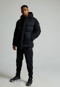 Svart pufferjakke med glidelåsfeste foran, polstret design og hette. Brukt med svarte joggebukser og sportssko med grå detaljer.