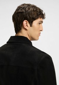 Svart mockajacka med en strukturerad krage, synliga sömmar och en subtil logodetalj på baksidan. Slät yta och kroppsnära design.