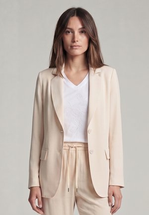 Frau trägt beige maßgeschneiderten Blazer über einem weißen bestickten V-Ausschnitt-Shirt mit passenden beigen Kordelzughosen, stehend vor hellgrauem Hintergrund.
