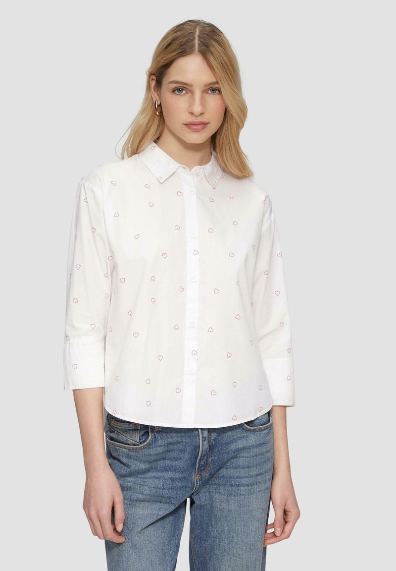 Chemise blanche à boutons avec des motifs de cœurs roses, manches longues et col. Associée avec un jean en denim bleu.