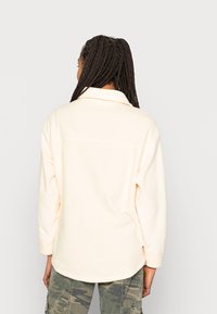 Camisola de fleece pêssego claro com colarinho, ombros caídos e mangas longas. Apresenta uma bainha arredondada e detalhe de yoke nas costas. Usada com calças de padrão camuflado.